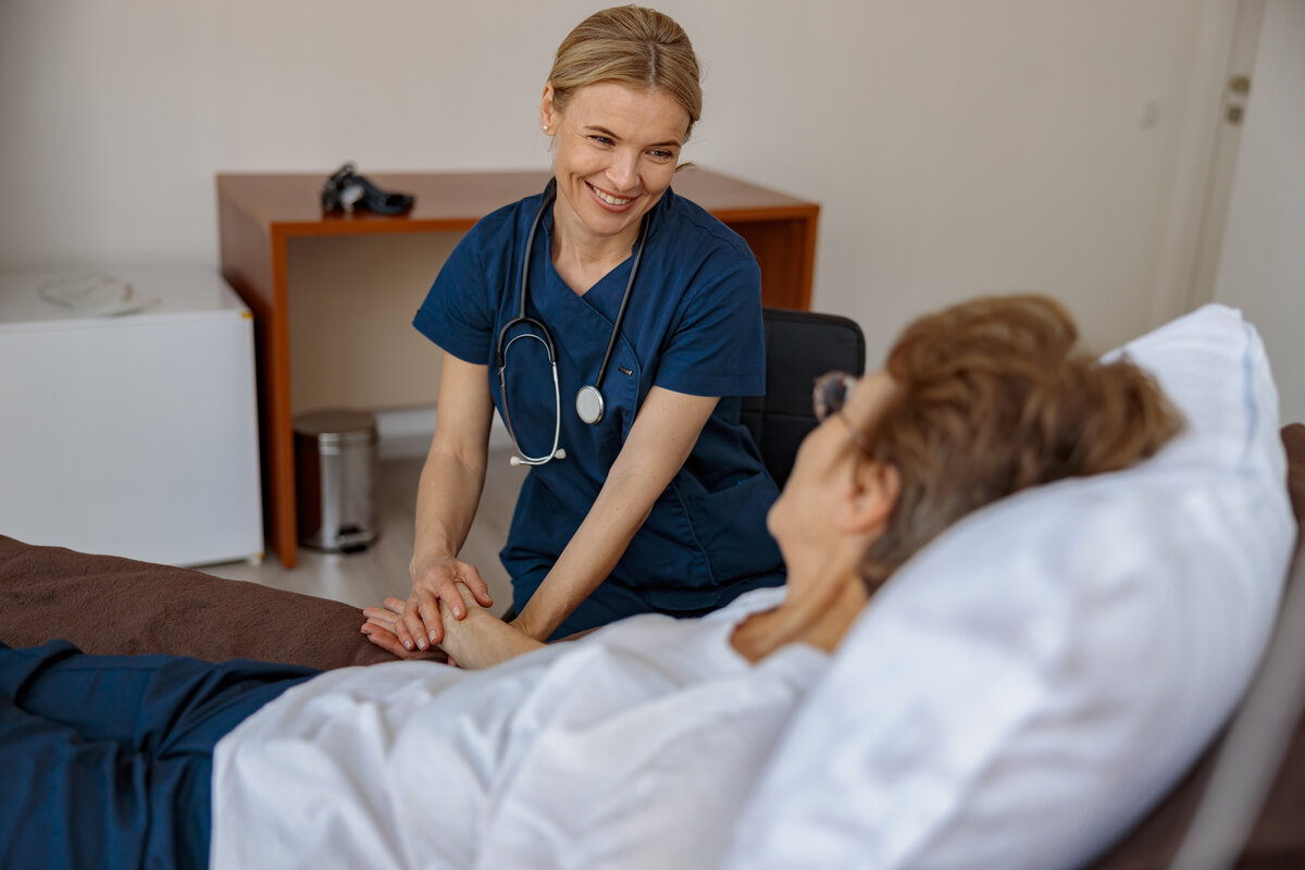 Blonde nurse in blue scrubs providing compassionate bedside care to patient